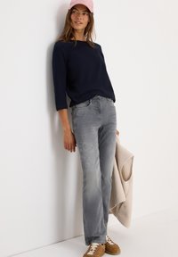Kvinna i en rosa keps, marinblå topp, grå jeans och bruna sneakers lutar sig mot en vit vägg, hållande en beige fleeceväst.