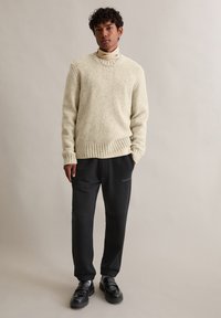 Suéter de malha texturizada em bege claro com bainha canelada, sobre um gola alta creme, combinado com calças pretas e mocassins pretos.