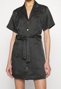 Robe en satin noir à manches courtes, avec boutons sur le devant et ceinture à la taille. Comprend une poche poitrine et des détails en métal doré. Texture lisse.