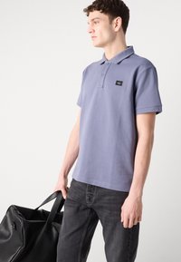 Polo à manches courtes en bleu clair, en tissu texturé, avec un col à deux boutons et un patch logo noir. Porté avec un jean sombre.