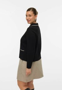 Kobieta z włosami upiętymi w kok, ubrana w czarną długą marynarkę, beżową spódnicę, czarne botki i wiszące kolczyki, uśmiechająca się do aparatu przez ramię.