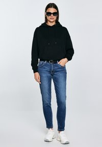 Fekete kapucnis pulóvert viselő nő, kék farmernadrág fekete övvel, fehér sportcipővel és fekete napszemüveggel, egyszerű fehér háttér előtt áll.