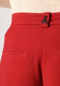 Pantalons taillés rouges dotés d'une texture lisse, d'un devant plat, de poches latérales et d'une fermeture en métal noir avec un détail décoratif.