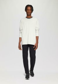 Lange mouwen lichtgrijs shirt met zwarte halslijn, gecombineerd met zwarte broek en zwarte sneakers, staande tegen een neutrale achtergrond.