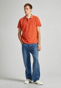 Oranžové polo tričko s krátkým rukávem, nošené s světle modrými rozšířenými džíny a bílými teniskami. Jednoduchý design, volný střih, bez viditelných vzorů.