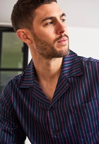 Camisa de pijama de rayas azul marino con rayas verticales rojas y blancas, mangas cortas, cuello de muesca y un bolsillo en el pecho. Textura de tela suave.
