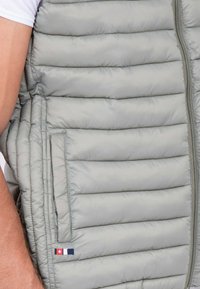 Gilet rembourré gris clair avec un matelassage horizontal, poches latérales et une fermeture éclair. Présente un logo de marque subtil en rouge et bleu sur le côté.