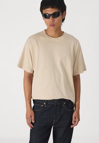 Camiseta de algodón beige con mangas cortas y cuello redondo. Combinada con pantalones vaqueros de denim azul oscuro con costuras naranjas y bolsillos.