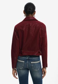Veste cropped en cuir bordeaux avec un col en V, des zips sur le côté et des pattes latérales ajustables, portée sur un jean bleu.