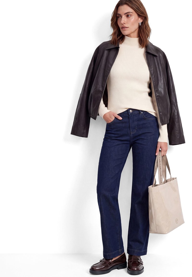 Vrouw draagt een donker leren jack over een crèmewitte trui, donkerblauwe spijkerbroek, bruine loafers, en houdt een beige tote tas vast.