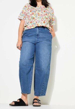 Person, der bærer en blomstret kortærmet skjorte, blå højhælede jeans med syninger på knæene og sorte slip-on sandaler med spænder.