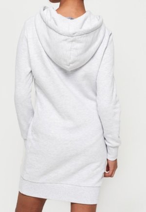 Osoba ubrana w sukienkę z kapturem i długim rękawem w jasnoszarym kolorze, przedstawiona od tyłu na jednolitym tle.