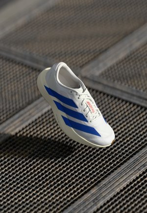 White athletic shoe with blue stripes, lightweight design, mesh upper, lace-up closure, and textured rubber sole, floating above a grid surface.