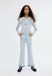 Light blue denim overalls with wide legs, featuring front pockets and adjustable straps, paired with a long-sleeve floral-printed shirt.