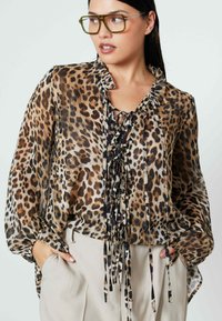 Luipaardprint blouse met een gerimpelde halslijn, sheer stof en strikdetails aan de voorkant. Lange, opgeblazen mouwen. Gecombineerd met lichtgekleurde broeken.