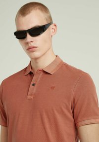 Polo shirt color ruggine con colletto classico, due bottoni e un piccolo logo. Occhiali da sole neri angolari con lenti scure. Texture liscia.