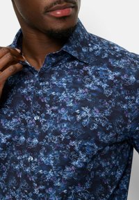 Blumenmusterhemd in Marineblau mit hellblauen und lila Akzenten. Verfügt über einen spitzen Kragen und Knopfverschluss.