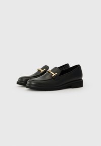 Mocassins en cuir noir avec de petits talons et des barres en métal doré sur le dessus, présentés de manière inclinée sur un fond blanc.
