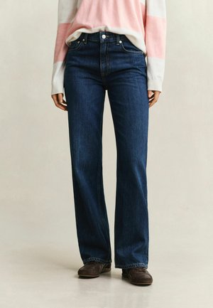 Personne portant un jean bleu foncé à jambe droite, un pull à manches longues rose et blanc à rayures et des chaussures marron foncé, debout sur un fond clair.