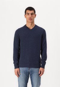 Mornarsko modra v-neck pulover iz mehkega pletenega materiala, z rebrastimi manšetami in robom, ter logotipom na spodnji levi strani. Oblečeno z modrimi kavbojkami.