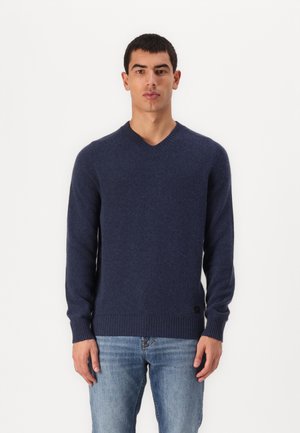 Pull en V bleu marine en tissu tricot doux, avec des poignets et un ourlet côtelés, et un détail de logo en bas à gauche. Porté avec un jean bleu.