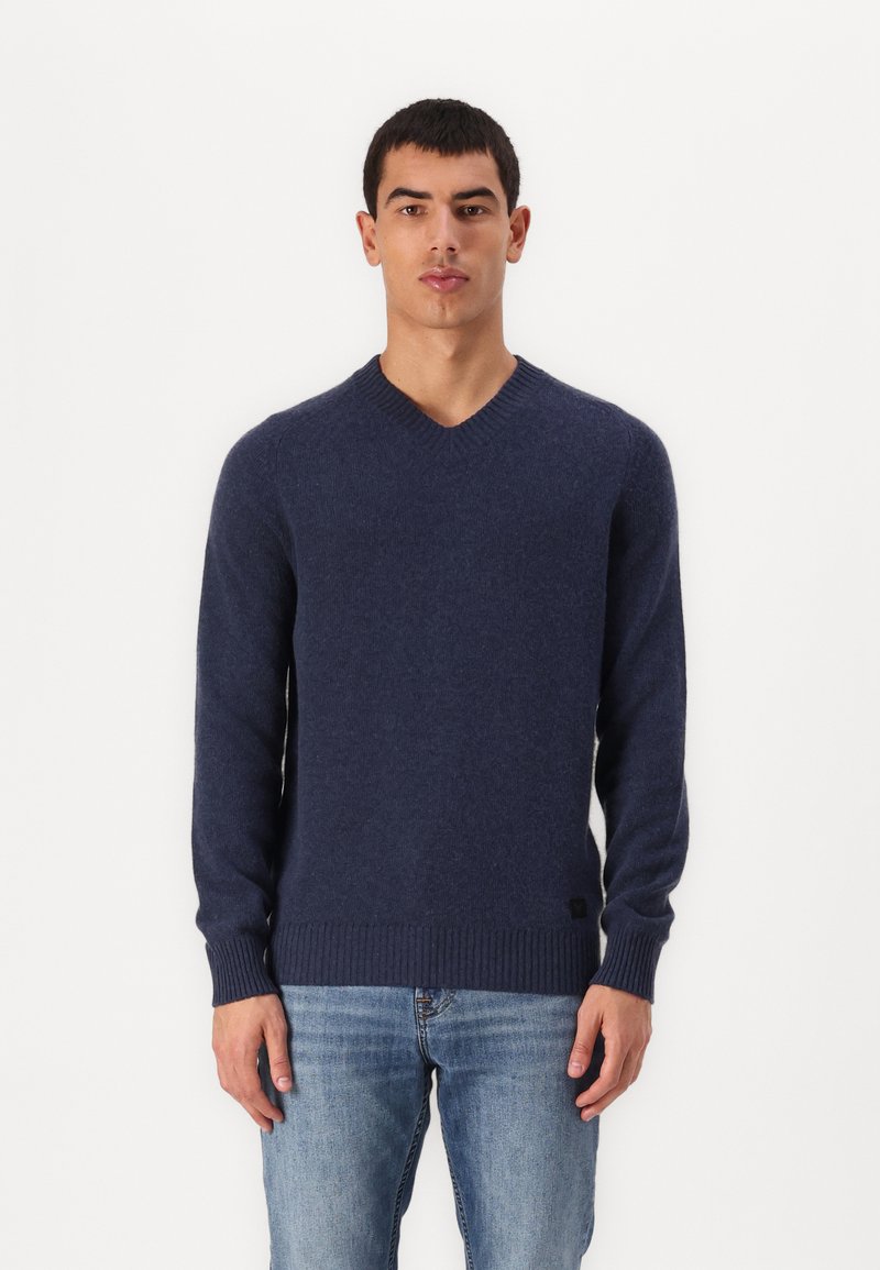 Mornarsko modra v-neck pulover iz mehkega pletenega materiala, z rebrastimi manšetami in robom, ter logotipom na spodnji levi strani. Oblečeno z modrimi kavbojkami.