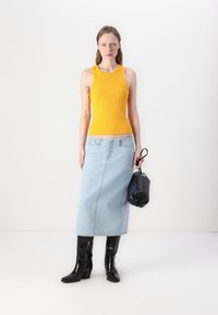Débardeur jaune côtelé, jupe midi en denim bleu clair, bottes noires montantes, et un petit sac à main noir. Fond blanc uni.