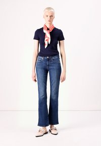 T-shirt marine, jeans évasés bleus et chaussures à enfiler beiges. Une écharpe à motifs rouges et blancs nouée autour du cou. Tissu lisse, coupe régulière.