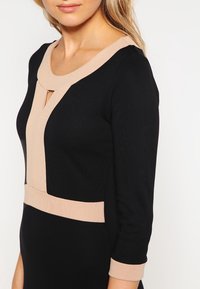 Czarna sukienka z beżowymi akcentami, z okrągłym dekoltem, detalem w kształcie dziurki i rękawami o długości trzech czwartych. Gładki materiał z kontrastującymi paskami.