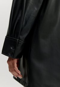 Schwarze Lederjacke mit glatter Textur. Lange Ärmel mit Knopfmanschette, die subtile Nähte und eine taillierte Passform zeigt.