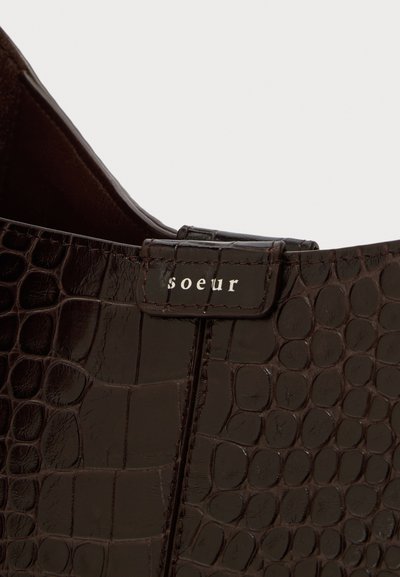 Γωνία τσάντας από συνθετικό δέρμα σε στυλ καφέ κροκό, με ραμμένη ετικέτα «soeur» στο καπάκι, πάνω σε φωτεινό φόντο.