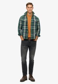 Camisa de franela en cuadros verde y azul marino, camiseta de cuello redondo naranja debajo, jeans grises, botas de cordones marrones y reloj de pulsera plateado.