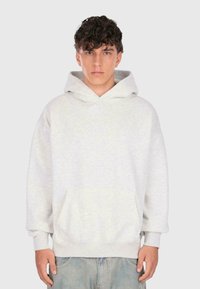 Un jeune homme aux cheveux bouclés porte un sweatshirt à capuche gris clair et un jean bleu, se tenant devant un fond gris clair uni.