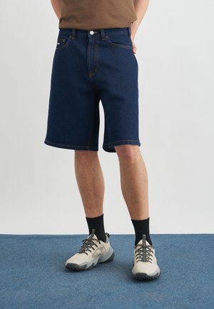 Homme portant un short en denim bleu foncé, des chaussettes noires et des baskets beiges, debout sur un tapis bleu contre un mur blanc uni.