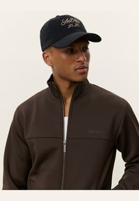 Καφέ zip-up μπουφάν με ψηλό γιακά, κατασκευασμένο από λείο υλικό. Μαύρο καπέλο με χρυσό κεντημένο λογότυπο "Les Deux Mr.", δημιουργώντας αντίθεση.