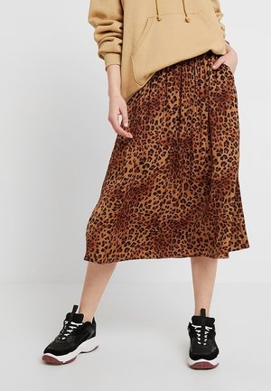 Person wearing a tan hoodie, leopard print midi skirt, and black sneakers with white soles standing with legs apart.