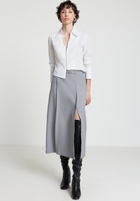 Weißes, zum Reißverschluss geöffnetes Hemd mit Kragen, kombiniert mit einem grauen Rock mit einem seitlichen Schlitz und schwarzen kniehohen Stiefeln mit Absatz. Glatter Stoff mit maßgeschneiderter Passform.