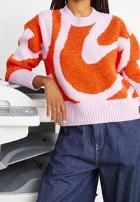 Pull tricoté coloré avec des motifs abstraits roses et oranges, doté d'un col rond côtelé. Associé à un pantalon en denim, sur un fond d'imprimante.