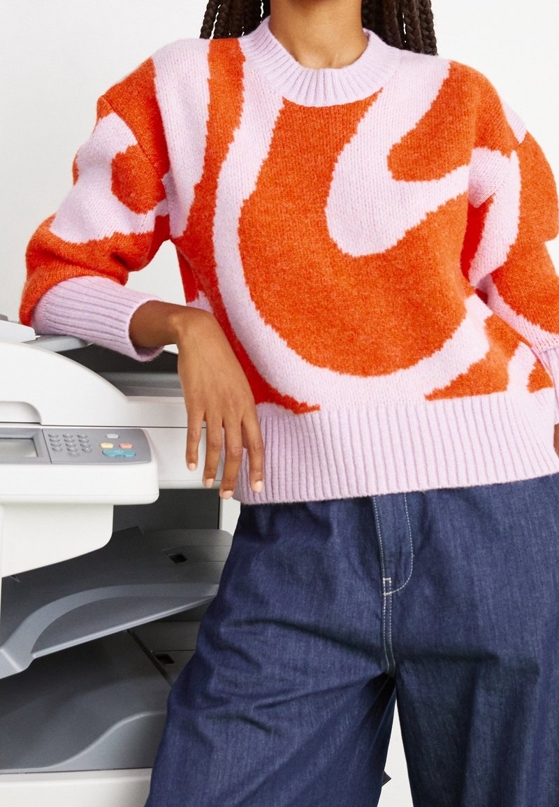 Pull tricoté coloré avec des motifs abstraits roses et oranges, doté d'un col rond côtelé. Associé à un pantalon en denim, sur un fond d'imprimante.