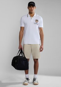 Polo bianco con accenti neri e dorati, logo sul petto, pantaloni corti beige, calze bianche e sneakers multicolori. Borsa a tracolla nera in mano.