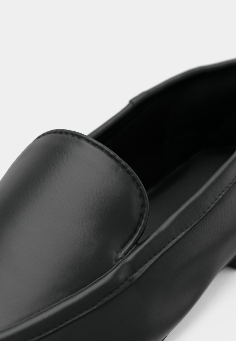 Chaussure noire en cuir à enfiler avec un bout arrondi et une finition lisse. Couture visible le long du bord supérieur. L'intérieur est élégant et minimaliste.