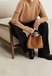 Sac à main en daim marron de forme arrondie avec des anses courtes, tenu par une personne portant un pull en maille texturée marron et un pantalon noir.