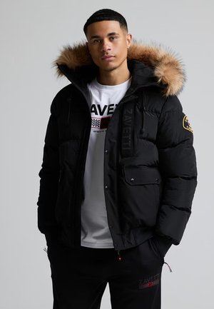 Un homme pose devant un fond gris uni. Il porte une doudoune noire à capuche bordée de fourrure, un T-shirt blanc à motif graphique et un pantalon noir.