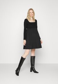 Vestido preto com um corpete ajustado em malha e uma saia evasê. Modelo usa botas pretas até ao joelho. Tecido suave com linhas limpas.