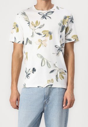 Homme portant une chemise blanche à manches courtes avec des imprimés floraux abstraits jaunes et gris, associée à un jean bleu clair, vue de face, debout.