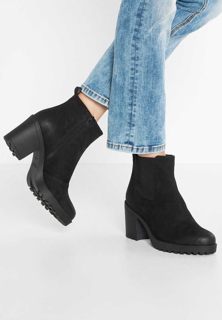 Bottes noires en daim avec talons épais et fermeture éclair latérale, portées avec un jean clair coupé court, sur un fond blanc.