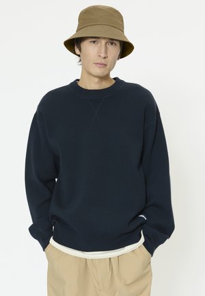 Marineblaues Sweatshirt mit Rundhalsausschnitt, überschnittenen Schultern und gerippten Bündchen, getragen über einem weißen Hemd. Ein brauner Fischerhut vervollständigt den Look.