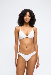 Γυναίκα με σγουρά μαλλιά που φοράει ένα λευκό σουτιέν και αντίστοιχο εσώρουχο Calvin Klein, στέκεται μπροστά σε ένα απλό φωτεινό φόντο.