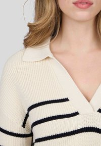 Pull en tricot crème avec des rayures bleu marine. Doté d'un col en V et d'un col. La texture est côtelée, mettant en valeur les couleurs contrastées.