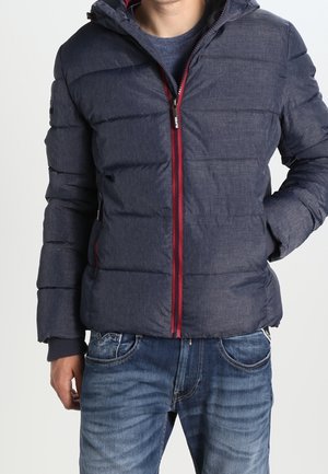 Homme portant une veste matelassée à capuche bleu marine avec une fermeture éclair rouge et un jean bleu, debout les mains dans les poches sur un fond uni.